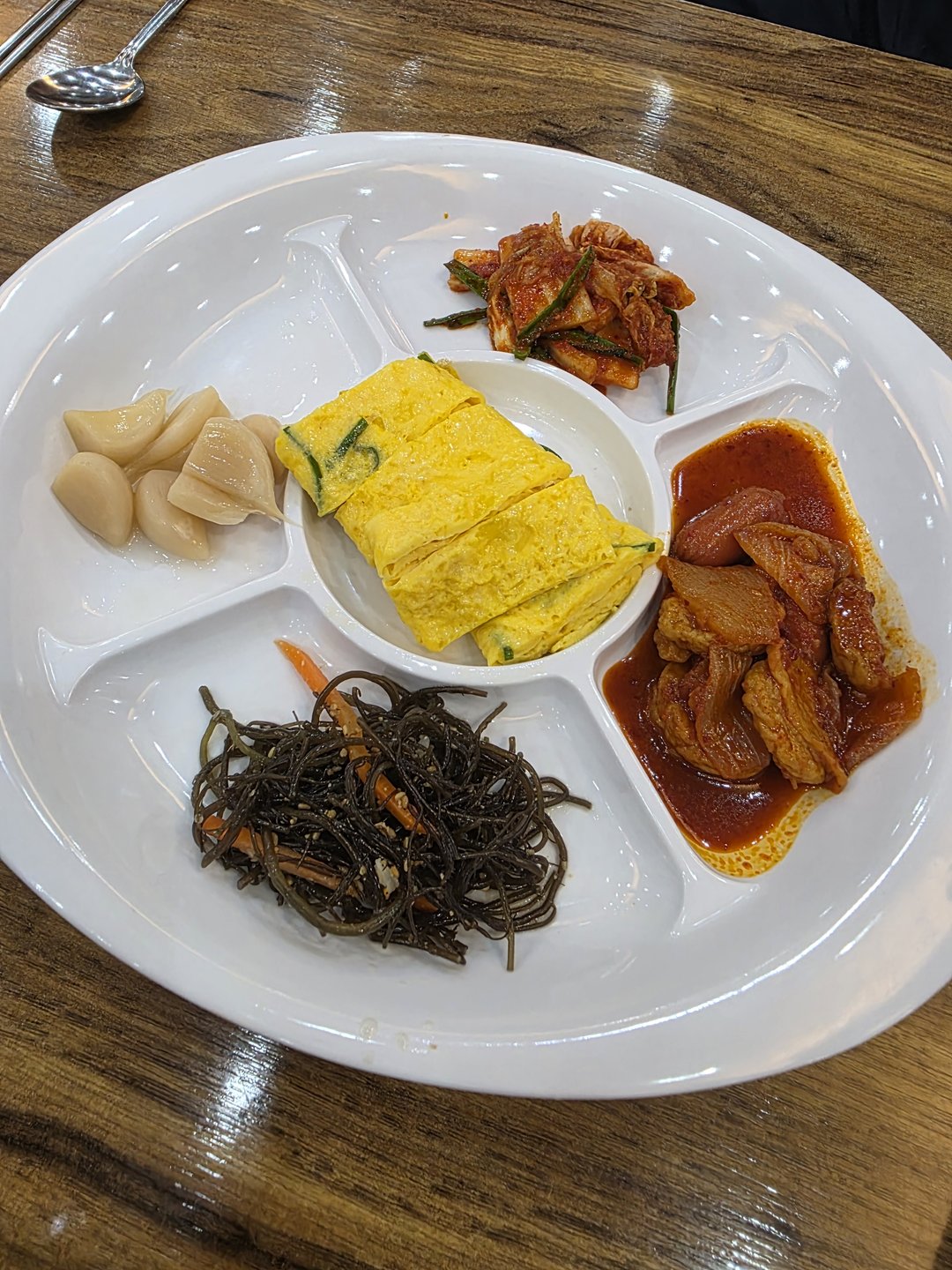 계란말이와 김치, 마늘장아찌, 오뎅볶음이 담긴 흰 반찬 접시