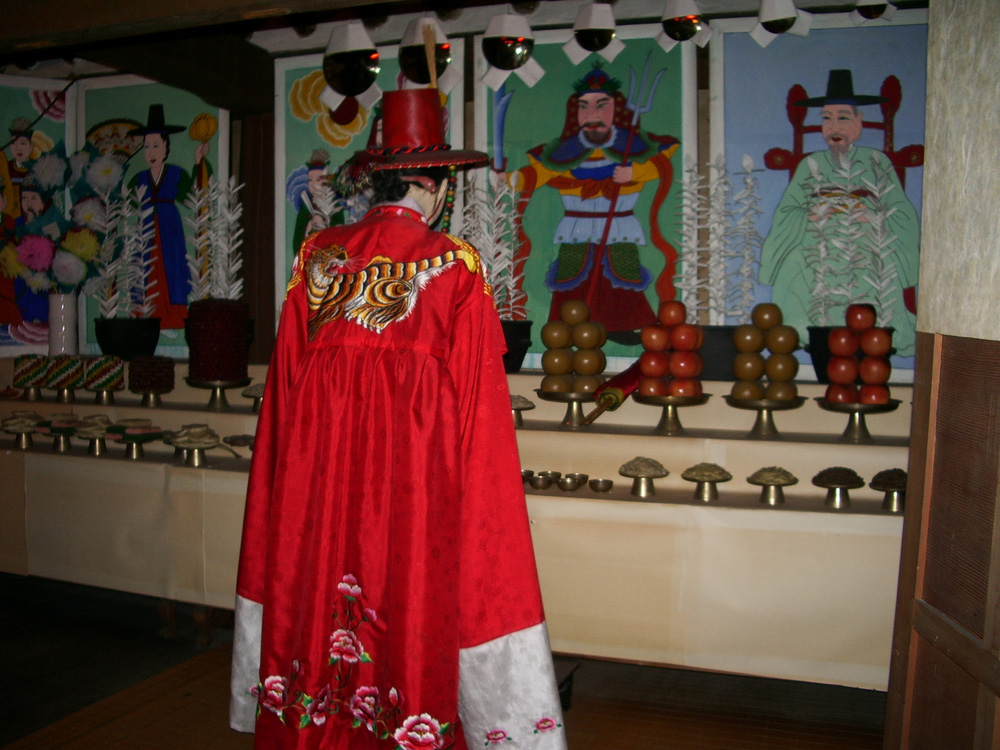 붉은 무복을 입은 무당이 무신도와 제단 앞에 서 있는 모습 / A Korean mudang in red ritual robes before a shamanic shrine