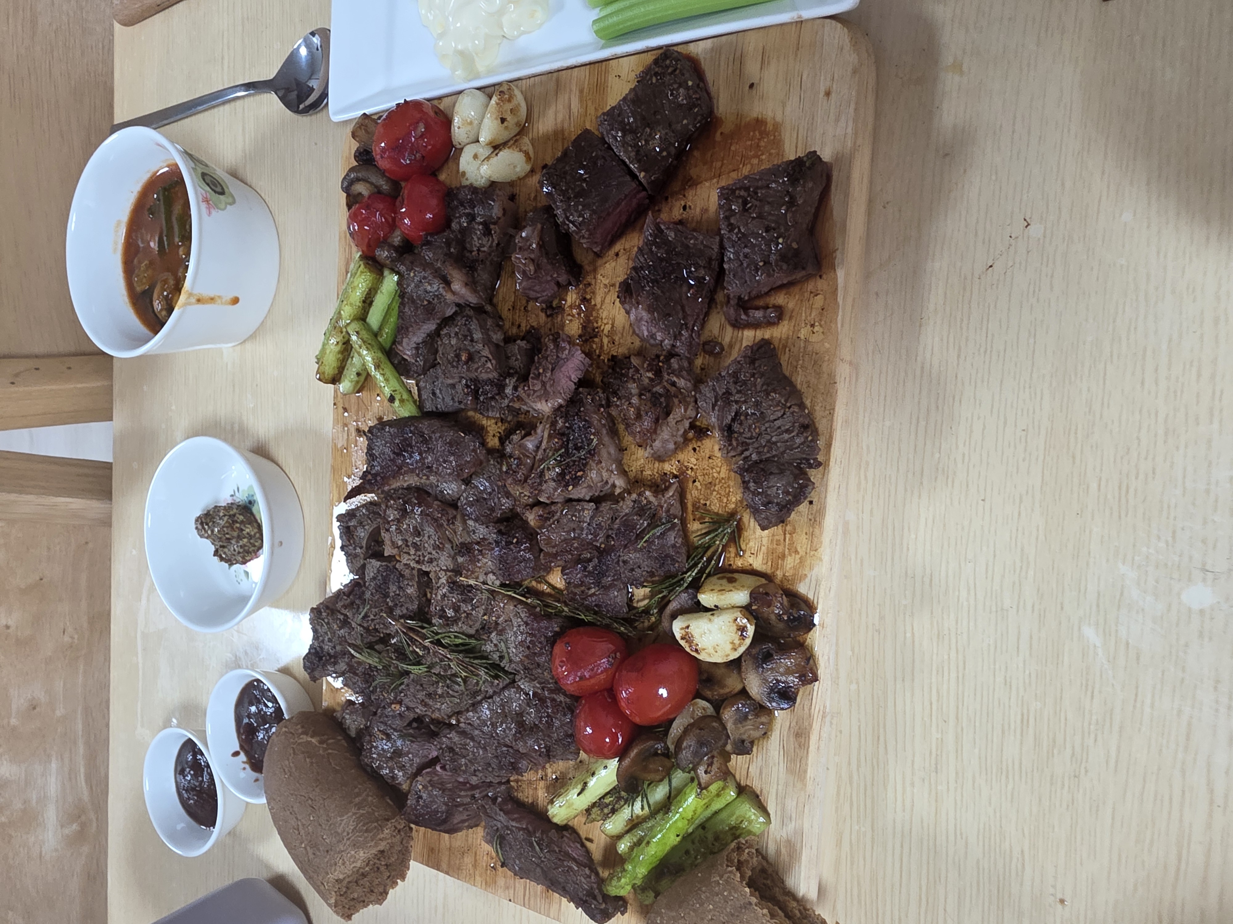 나무 도마에 먹기 좋게 썰어 올린 스테이크와 구운 방울토마토, 마늘, 아스파라거스, 로즈마리, 옆에는 소스 종지 세 개와 셀러리 접시