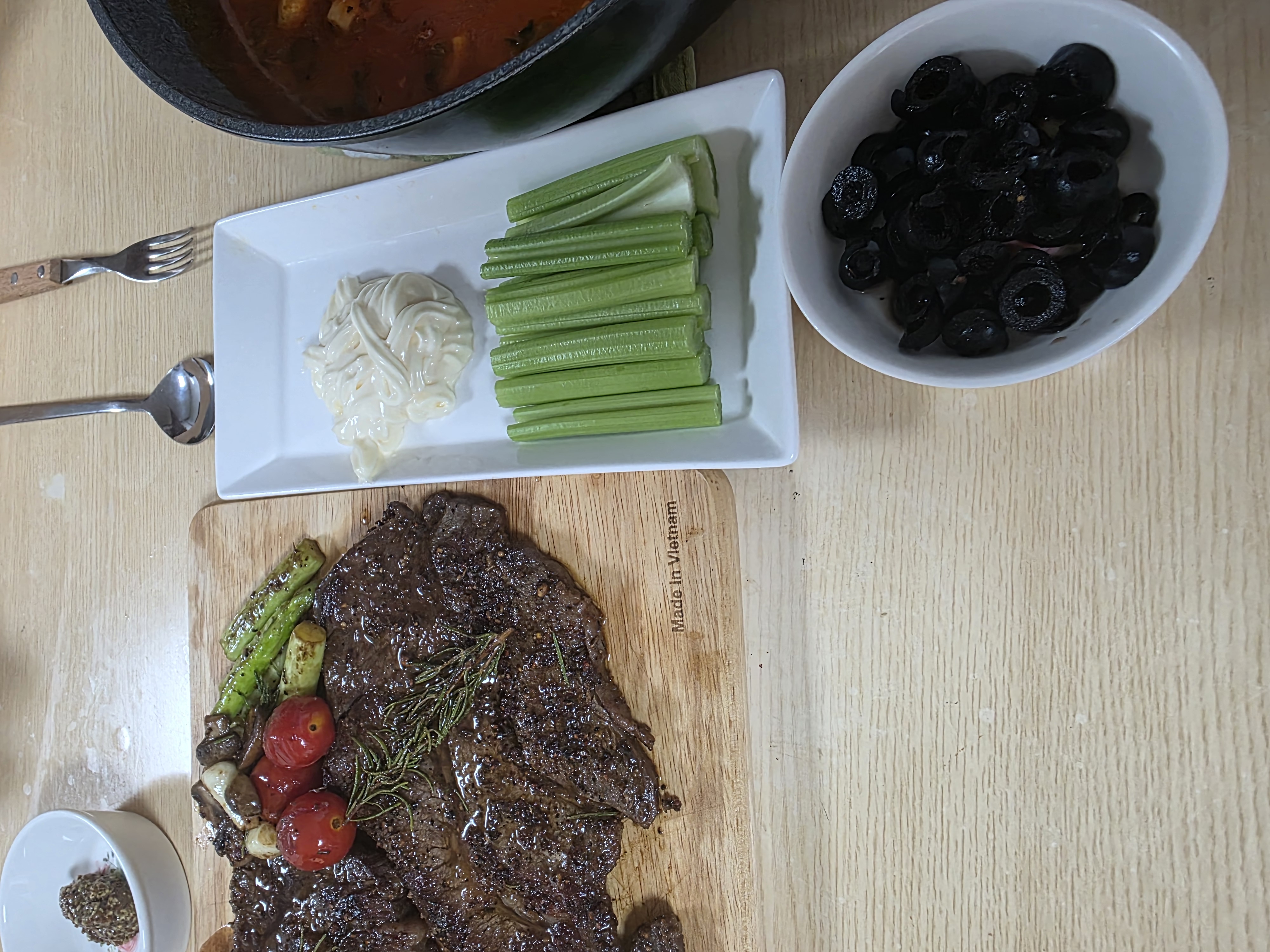 식탁에 차린 저녁, 도마 위에 통째로 구운 스테이크와 로즈마리, 옆에는 셀러리와 마요네즈 접시, 검은 올리브 한 그릇, 토마토 스튜가 담긴 무쇠 냄비