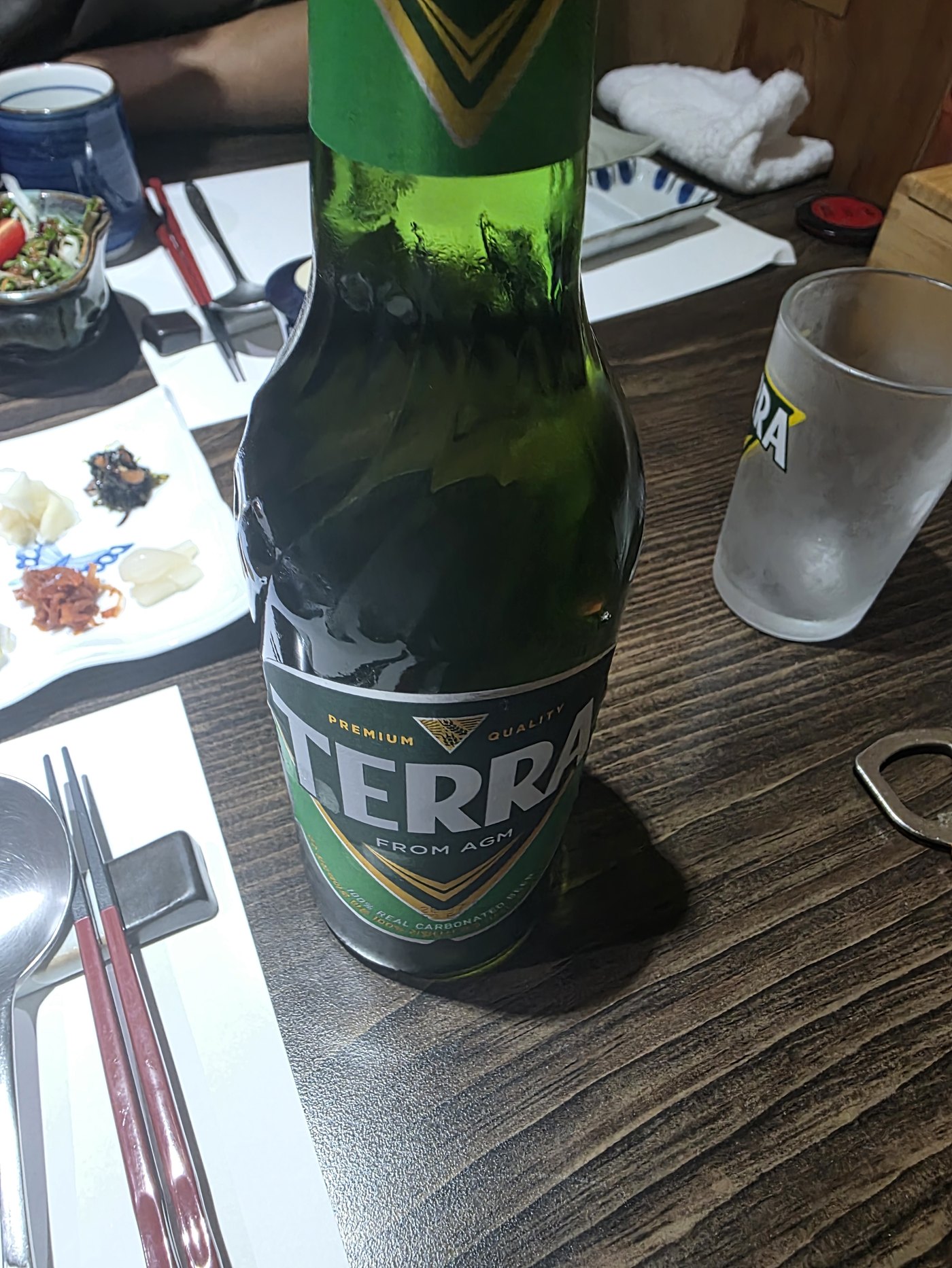 나무 테이블에 놓인 초록색 병맥주와 성에 낀 유리잔, 맞은편에 앉은 친구의 팔이 살짝 보이고 마늘장아찌와 젓갈이 담긴 흰 접시가 놓여 있다