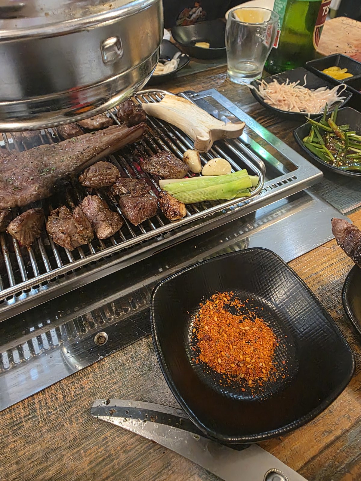 같은 석쇠 위 양갈비 옆으로 새송이버섯과 마늘 대파가 구워지고 있고 테이블 한쪽에 초록색 병맥주와 숙주 파절이 반찬이 놓여 있다
